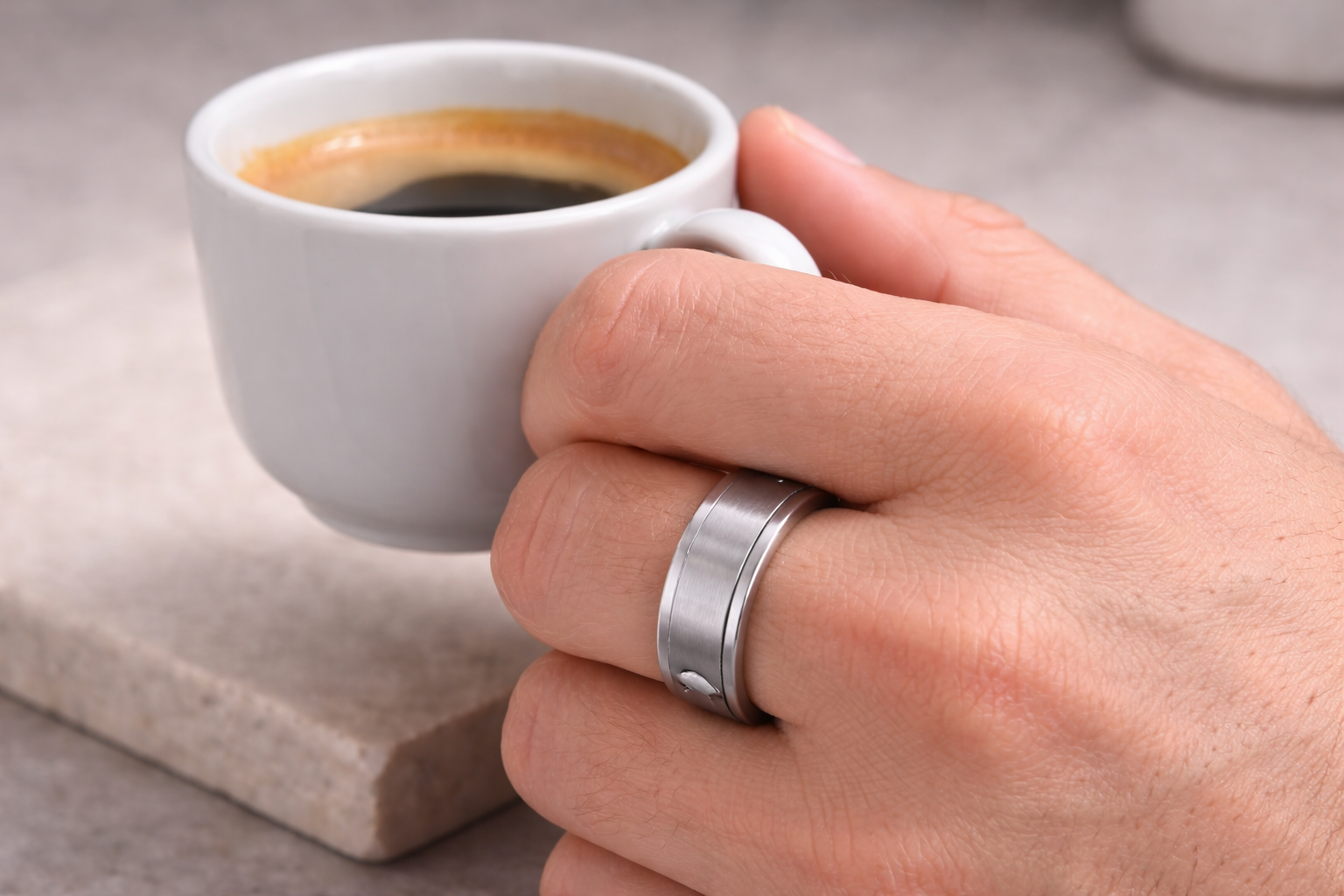 Hand holding a small white cup of coffee with a silver ring on a neutral background