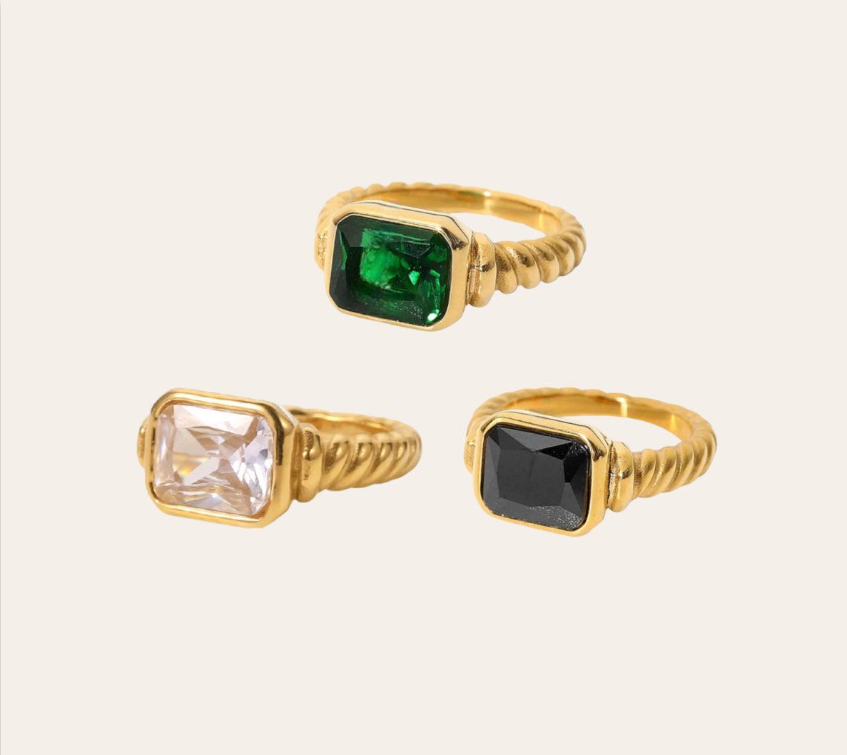 A set of three rings with square-shaped stones, the stones are green, clear, and black, all set in gold-colored bands.